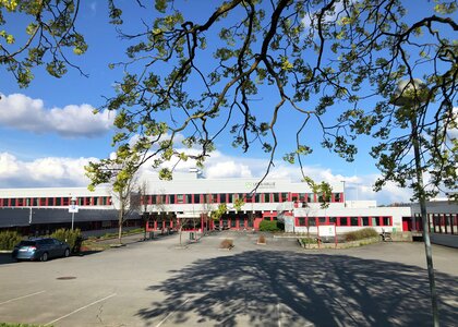 Grått skolebygg i to etasjer med røde vinduskarmer,  og en stor asfaltert skoleplass foran. - Klikk for stort bilde